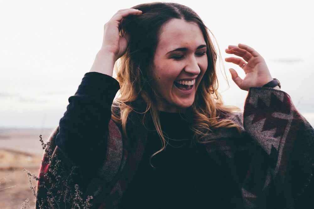 a white woman in her 20s with brown and bleached blonde hair stands in a field wearing a warm poncho. she laughs with her eyes closed, running one hand through her hair.