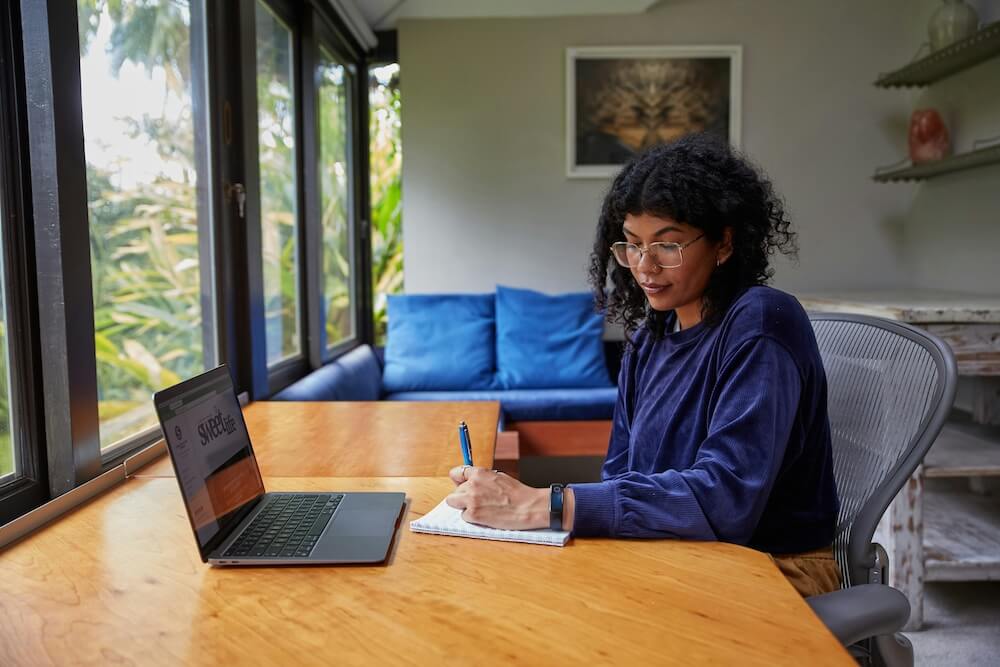 a feminine-presenting person of color with curly dark brown hair and glasses sits at a large wooden table in a comfortable living room with white walls, a painting, a blue couch, and floor-to-ceiling windows. They hold a pen in their left hand and is writing in a notebook. There's a laptop on the table in front of them. 