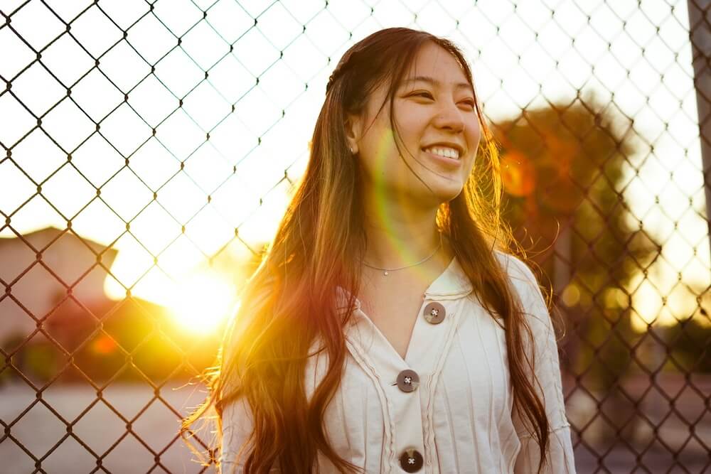 A young millennial Asian woman with long brown hair and a cream-colored shirt with large wood buttons stands in front of a chain-link fence. The sun is setting in the background behind her; she smiles as she looks off to the side, away from the viewer.