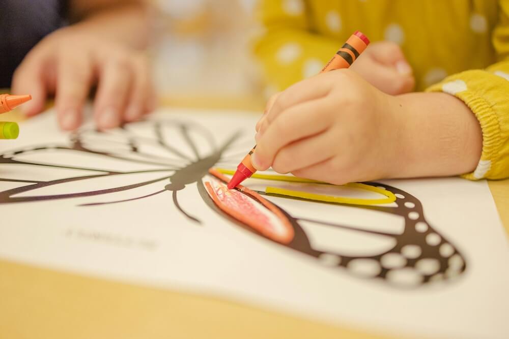 The hands of a child in a yellow long-sleeve top with white polka dots. They hold an orange crayon and are coloring in a picture of a butterfly. 