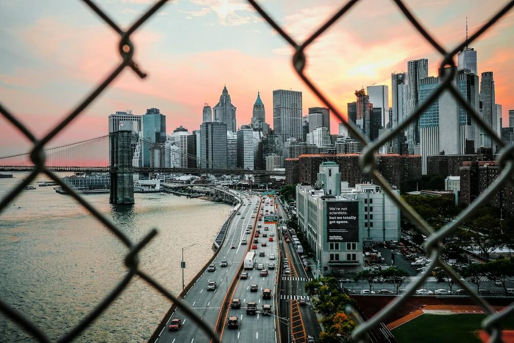 A sunrise or sunset behind a lakeside cityscape. In the foreground is a chainlink fence with a large hole cut through it.
