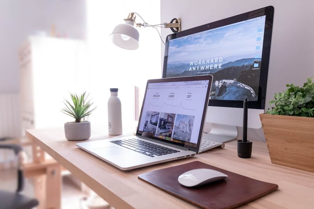 A brightly lit office space with a modern light-colored wood desk. A couple of small plants sit on the desk, along with an Apple laptop and desktop computer, cognac leather mousepad, and sleek white mouse.