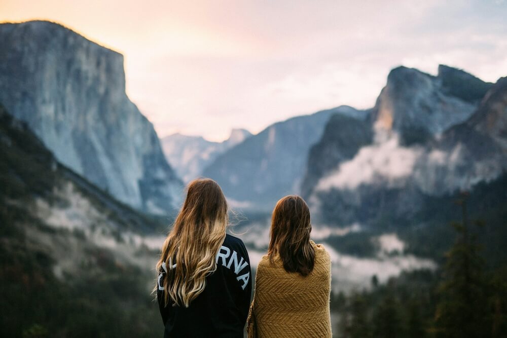 Two women with dark blonde hair have their backs to our view, watching mist in the distant mountains at dawn or dusk. One has longer hair to mid-back with highlights and waves; she wears a black sweater that reads CALIFORNIA in all caps across the back. The others' hair is straighter and just below the shoulders; she wears a mustard-yellow blanket wrapped around her shoulders.