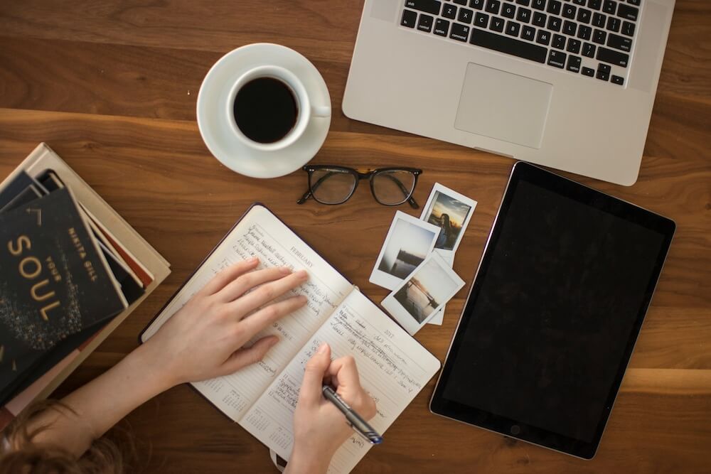 Feminine hands writing in a daily planner on a woodgrain desk, which also has eyeglasses, photographs, books, a cup of coffee, and table, and a laptop on it