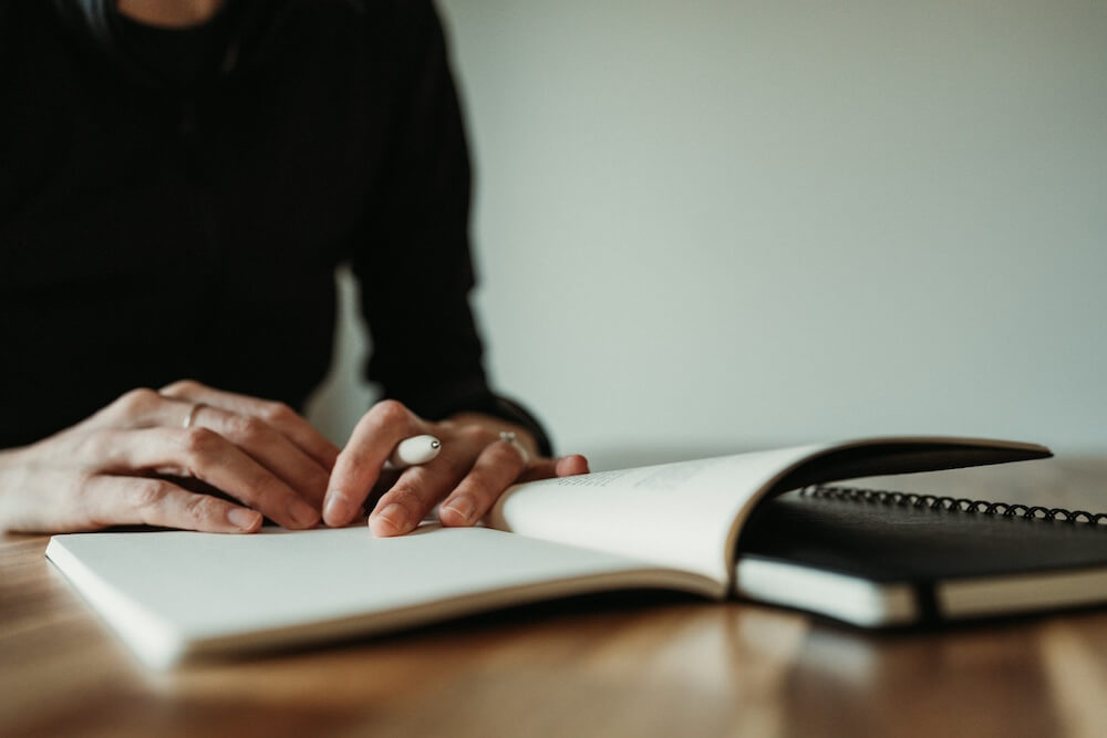 The hands of a woman wearing a black sweater and a few simple rings, holding a white pen. A notebook is open on the table in front of her, but her hands rest on the page as though she's deep in thought before writing.