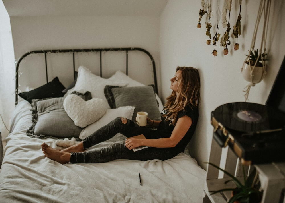 A white woman with brown and blonde hair sits on a cozy bed with neutral linens. She wears a black t-shirt and ripped dark gray jeans. She looks melancholy and is passively gazing out a window, holding a white coffee mug in one hand while resting the other on a notebook that sits on top of her outstretched leg.