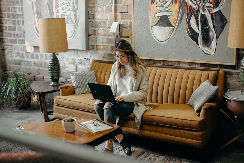 A young Asian woman with blonde highlights wears a cream-colored sweater and ripped black jeans. She sits on an orange couch in a coffee shop, with a laptop in her lap. She's typing and looking at the screen.