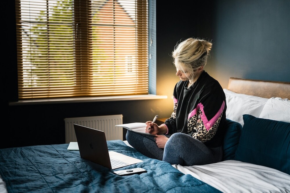 Blonde woman with undercut wears black, pink, and leopard-print sweater and sits cross-legged on a bed. She's writing in a journal and also has a laptop in front of her.