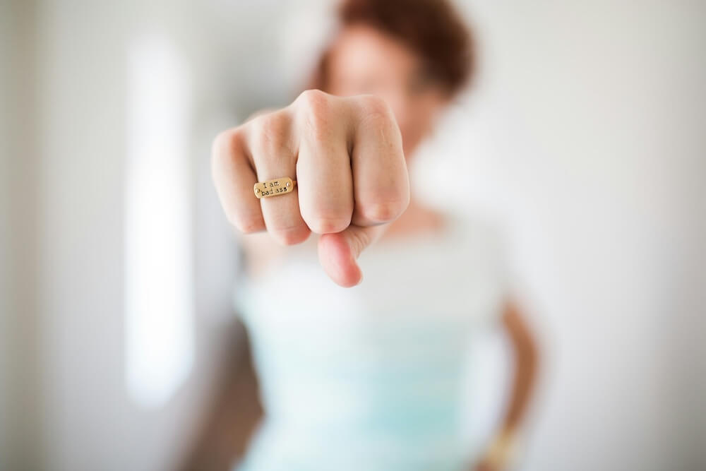 A close-up image of a white woman's fist; she wears a gold ring stamped with the words "i am bad ass". In the background, we see she has red hair and wears a white t-shirt, but she's completely out of focus except for her outstretched fist.
