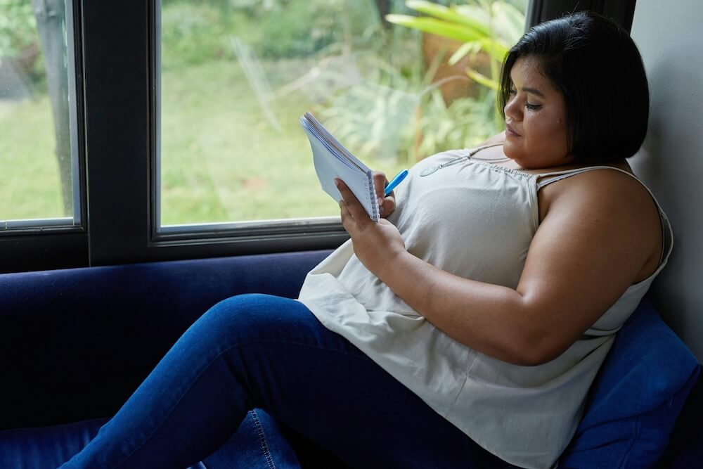 A plus-sized young woman of color with shoulder-length dark brown hair wears a white tank top and dark blue jeans. She sits on a daybed by window and writes in a journal.