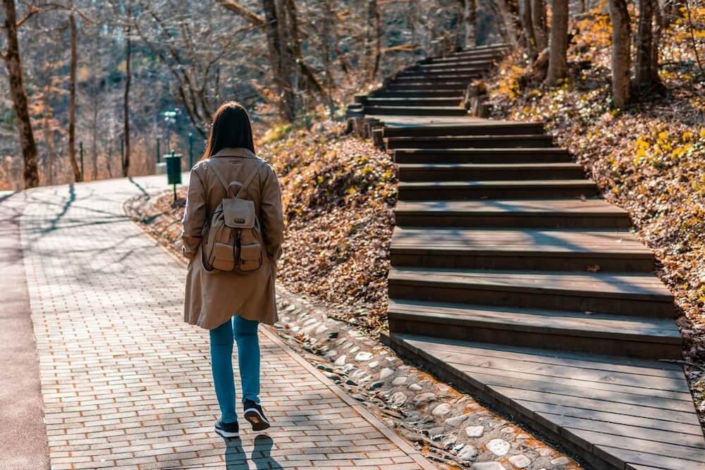 Woman with a beige coat and jeans wearing a leather backpack walks down a brick pathway; to her right is a set of stairs, representing a fork in the road in a career move or other decision
