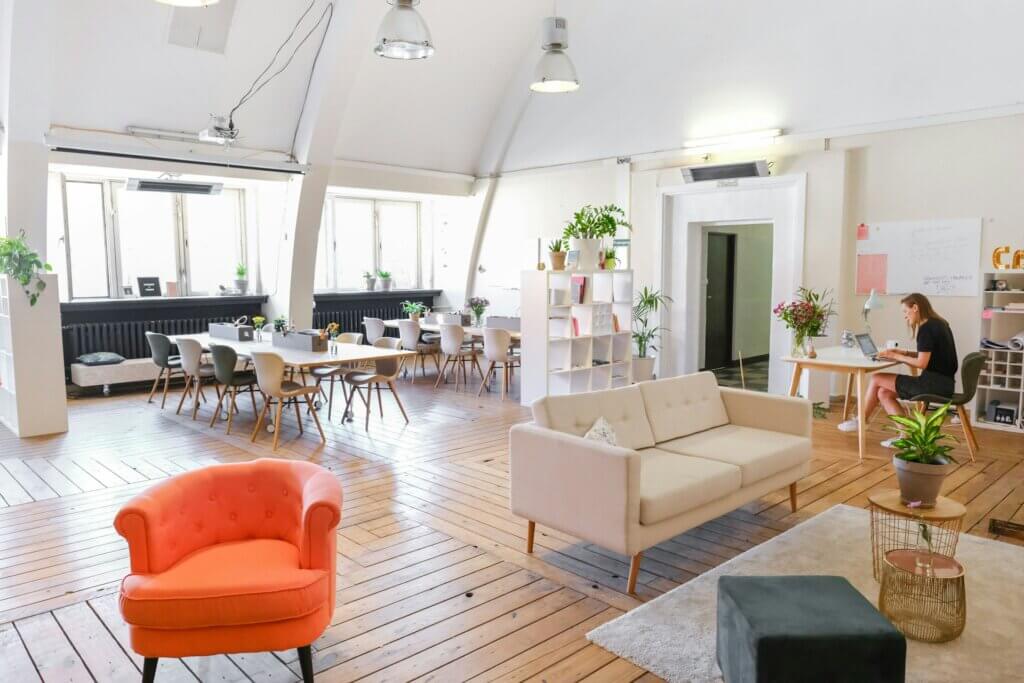 Large, bright office space with white walls and light wooden floors. Several desks and seating areas are scattered in a coworking space. One white woman sits alone at a desk, concentrating on her work. No one else is in the room.