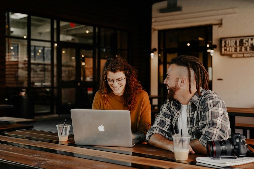 A young white woman with curly hair dyed red and young white man with brown-red dreadlocks and beard sit at a high-top table in a coffee shop. Both are laughing and the woman is looking down at her laptop. They both have iced coffees, and the man has a digital SLR camera nearby.