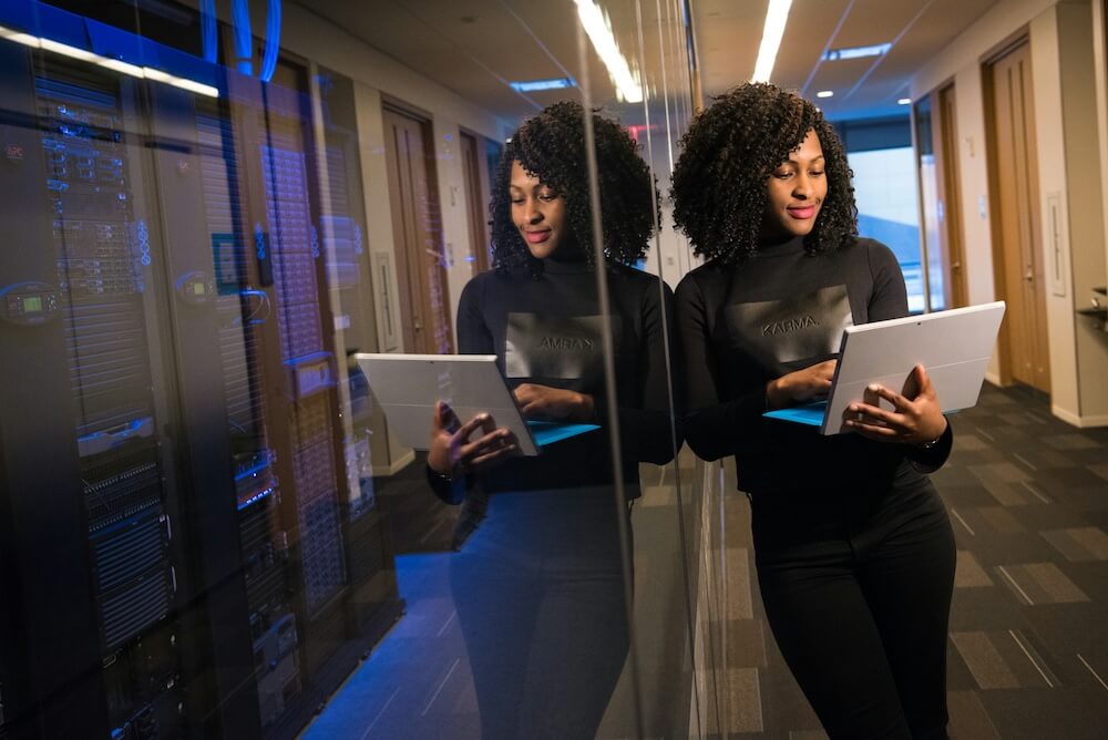 a Black woman with shoulder-length natural curly hair wears a black longsleeve shirt reading "KARMA" and black jeans. she stands leaning against the glass of a server room in an office setting. she holds a tablet and looks at the screen with a slight smile.