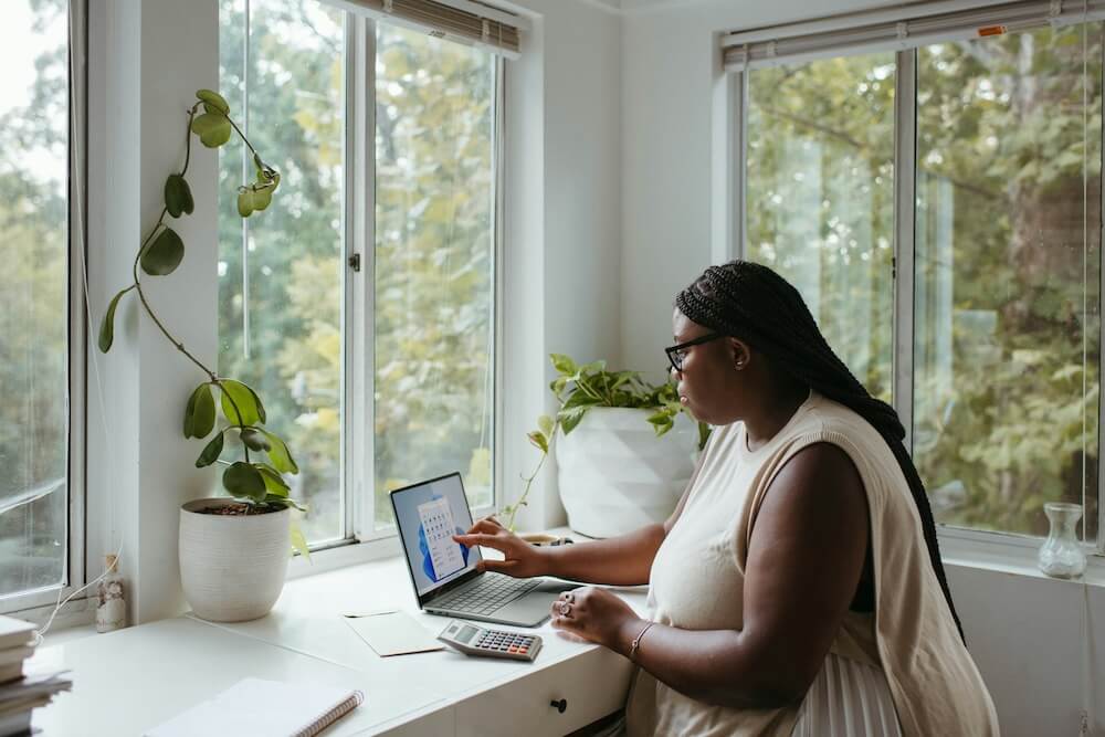 black woman with long braids wears cream-colored sleeveless dress and sits in a bright white office with multiple windows and green trees outside. she's working on a laptop.