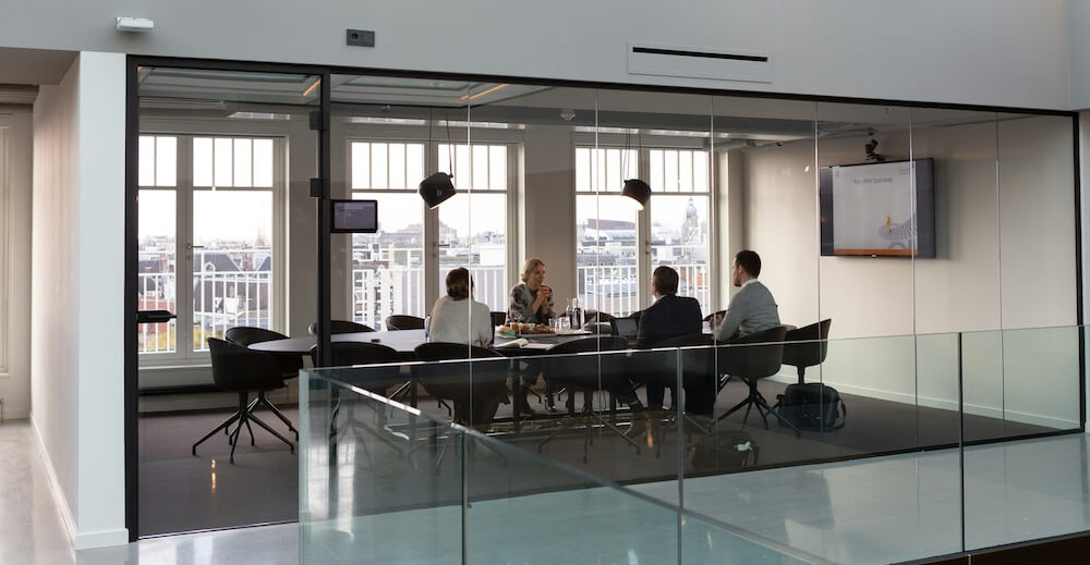the exterior of a glass conference room in a modern office building. inside, four people are meeting around a conference table.