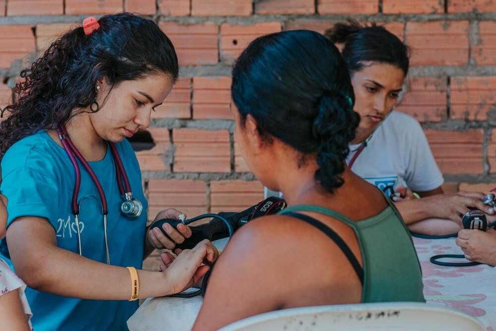 at an outdoor clinic in front of a brick wall, a young Latina woman in nurse uniform checks the pulse of a woman. in the background, another nurse does the same with another patient who's out of frame.