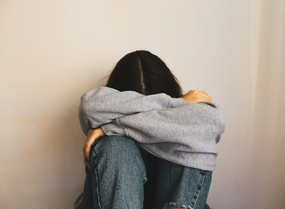 a person with long black hair parted in the middle sits on the floor, wearing a gray sweatshirt and jeans. they lay their arms across the top of their knees, and lay their head in their arms. their face isn't visible, but the vibe is frustrated and sad.