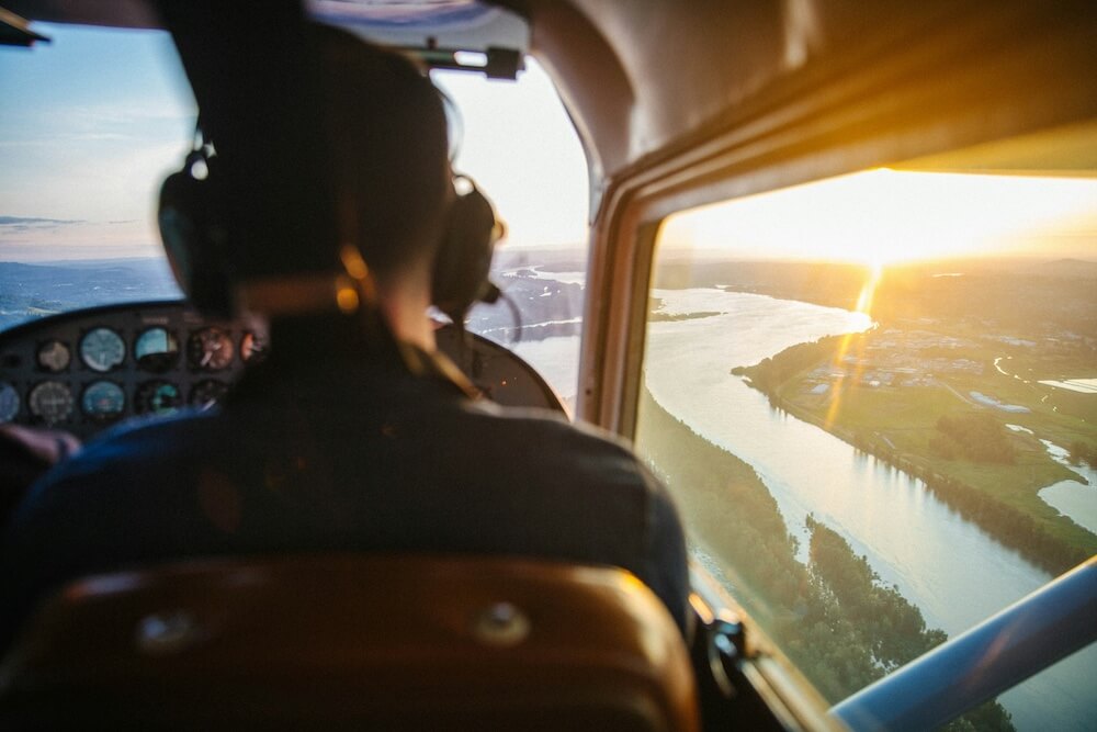 the back of a pilot's head with a view out the window of the plane as it flies over a river. the sun beams brightly on the horizon.