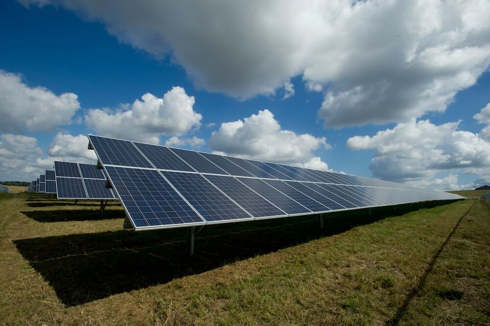 massive solar panels installed on the ground in a large grassy field. the blue sky and white fluffy clouds are above