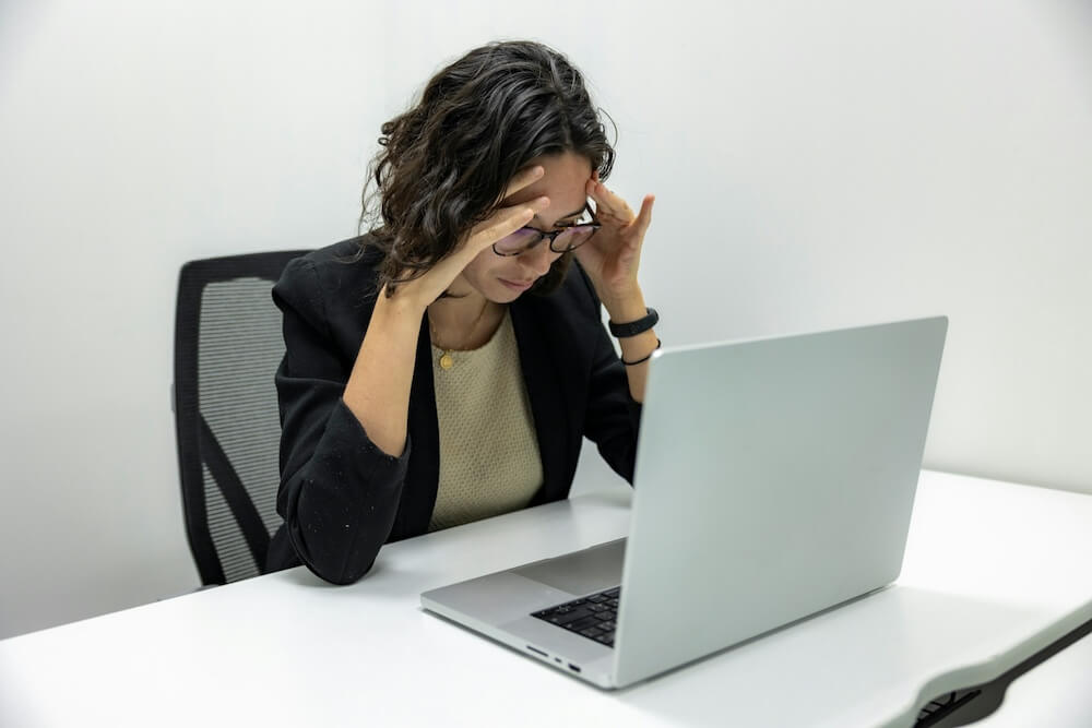 woman with short wavy brown hair sits at a white desk with hands raised to her temple. she rests her elbows on the desk and looks frustrated while staring at her laptop keyboard.