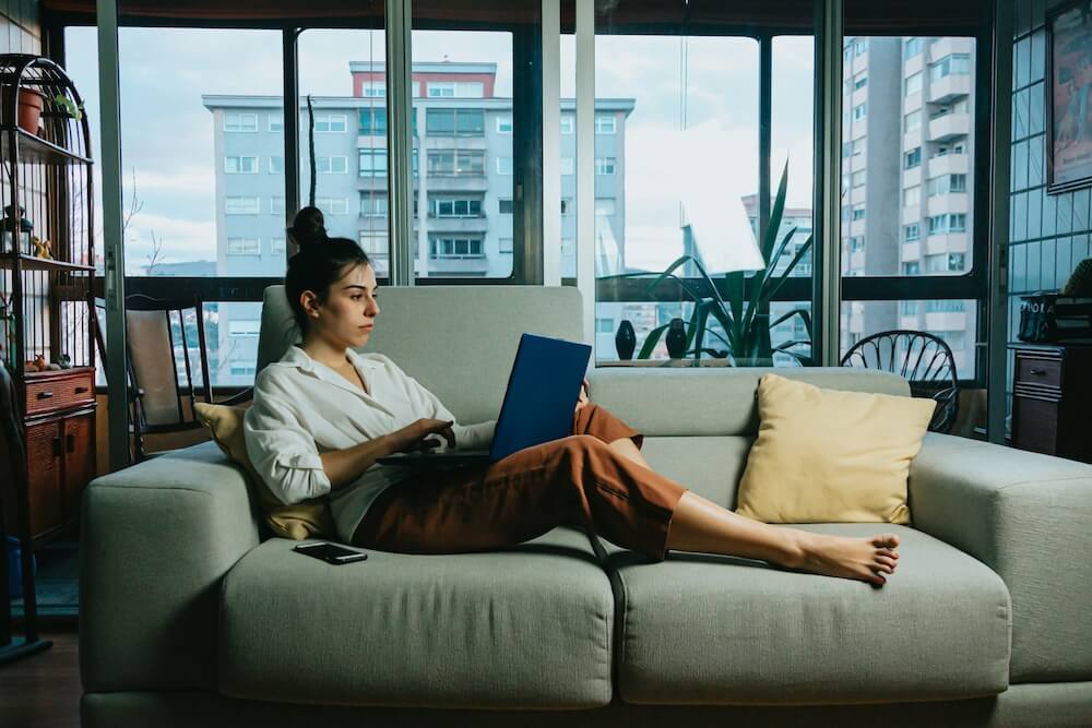 a woman of color with black hair wears a white button-down shirt and rust-colored cropped pants. she lounges on a light grey loveseat in an apartment, browsing on a laptop she has propped on one outstretched leg.