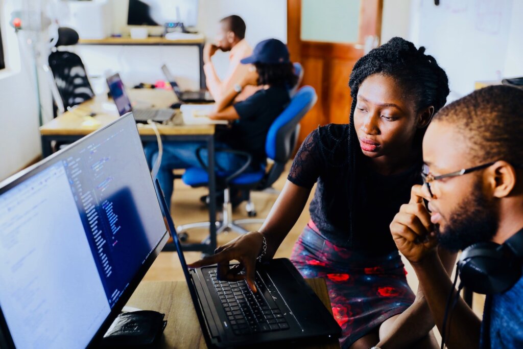 Black feminine-presenting person points to something on a laptop screen, as though showing the Black masculine-presenting colleague or student something to pay attention to. A larger computer monitor is visible with some code on it. In the background, 2 other people work at laptops, indicating a school or group study setting. Know what kind of education or training is required for your career change in late 30s.
