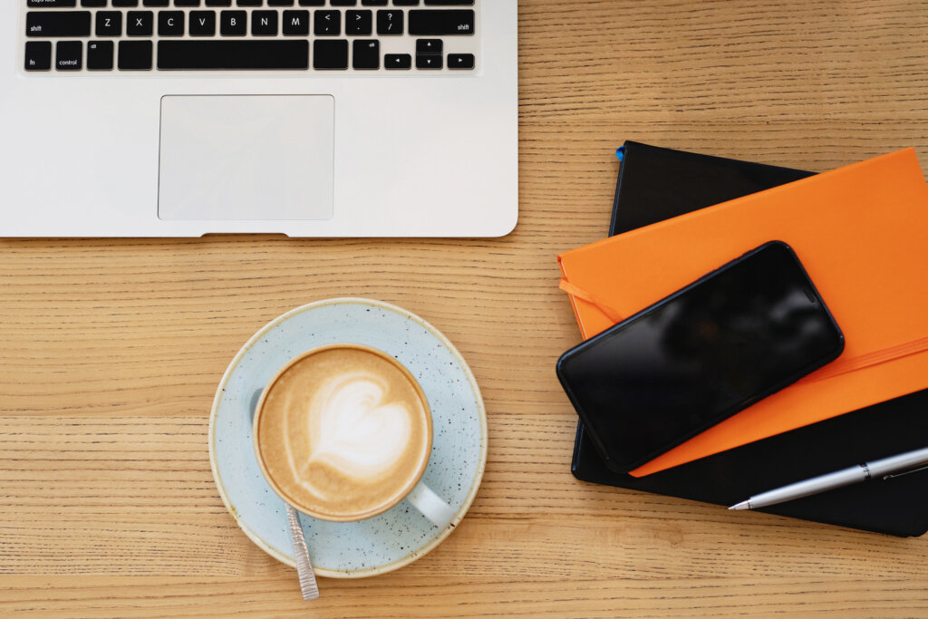 Latte with heart latte art in a mug and saucer, on a woodgrain table top with a laptop, black phone, black notebook and orange notebook with pen. You have to treat yourself when updating your resume!
