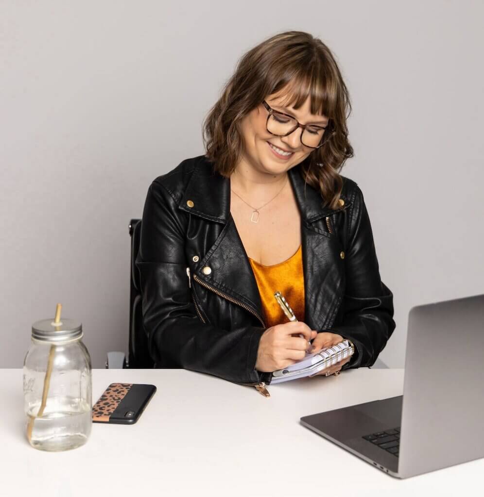 Coach Lucy Todd smiling with head tilted, wearing a black motorcycle jacket with rose gold accents, a metallic orange shirt, a silver pendant necklace, and brown glasses. She's writing in a small notepad as she sits at a white desk with gray laptop, mason jar filled with water, and phone with a black and leopard-print case.