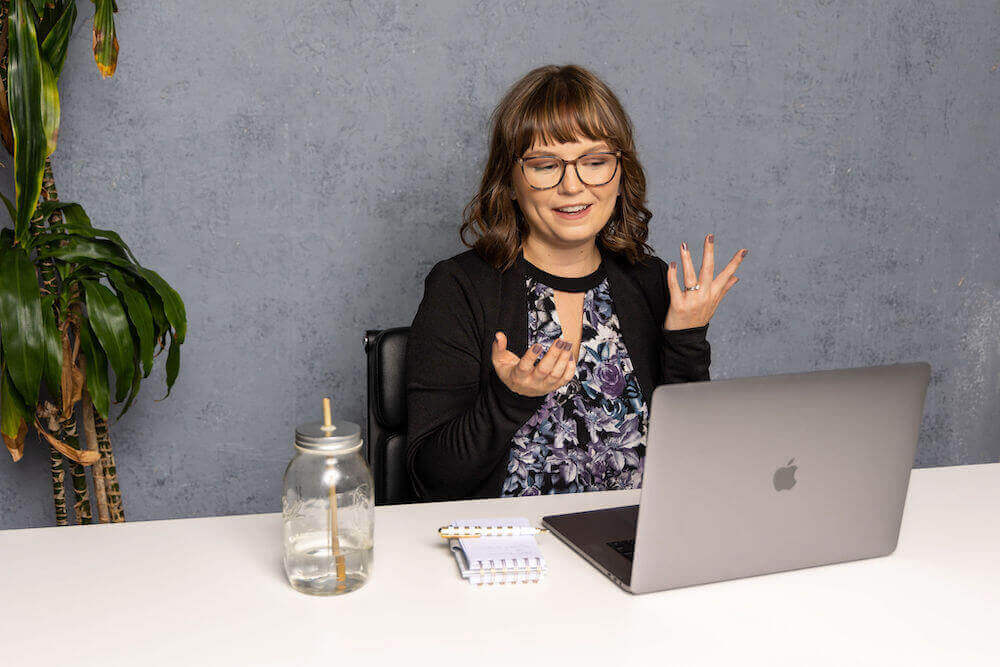 Lucy sitting in front of a gray backdrop at a white table. She appears to be on a video coaching session with a career coaching client, as she's mid-sentence and gesturing with her hands in the air while looking at her MacBook screen. She wears a purple floral top and black blazer.