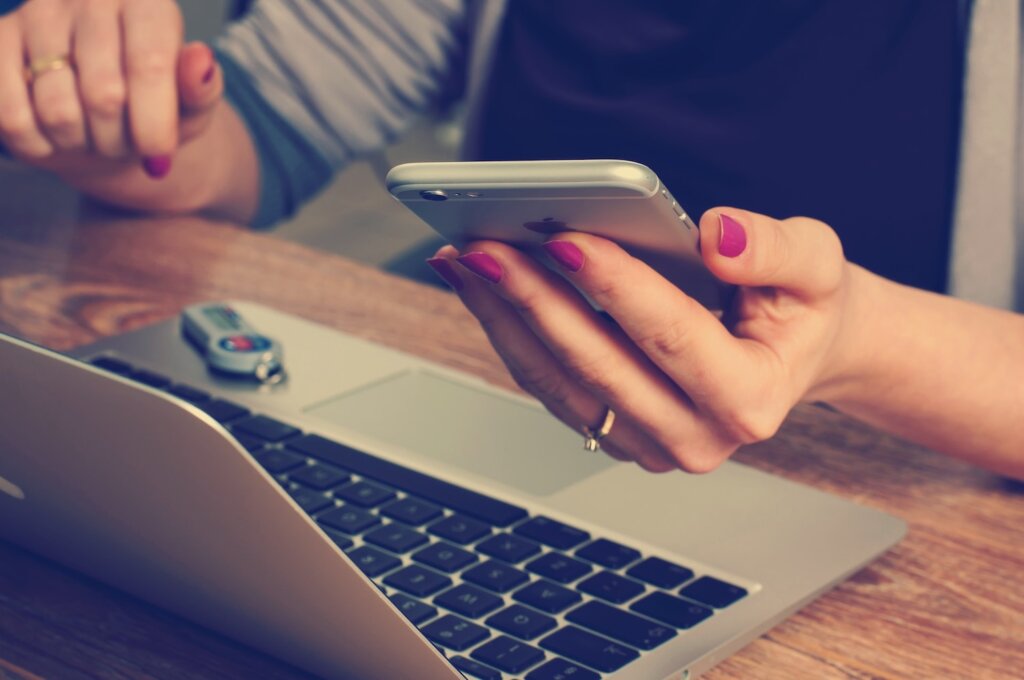 Feminine-presenting hands with pink nail polish and gold gemstone ring. One hand holds an Apple phone while the other hovers near a Macbook keyboard, which sits on a woodgrain counter.