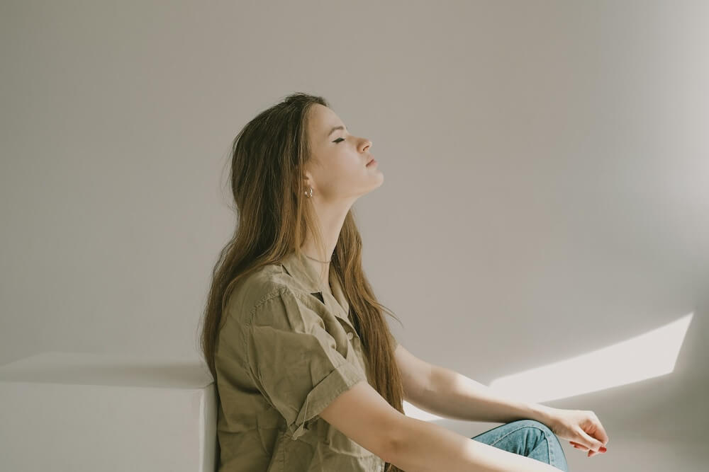 a white feminine-presenting person with long light brown hair wears a short-sleeved khaki button-down shirt and sits on the floor with eyes closed and head tilted up, as though meditating. she's in an all-white room with some sun shining in.