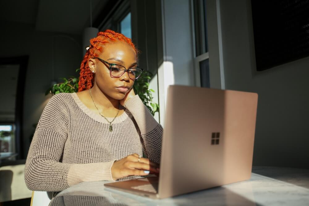 A young Black woman with bright reddish-orange braids, purple glasses, a pendant necklace, and a brown thermal top sits at a round marble table in a dark room, sunlight shining on her and the table. She props one elbow on the table and rests her face in that hand; the other hand hovers above the keyboard of a rose gold Surface laptop. She's ready to click on something with the trackpad.