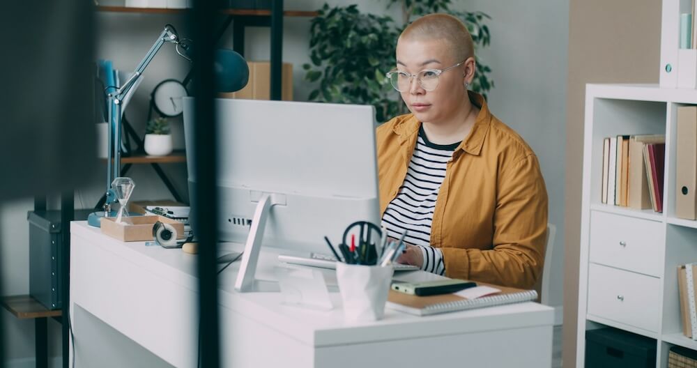 An Asian person with bleached buzzcut wears feminine vintage clear glasses, a black-and-white striped t-shirt, and unbuttoned mustard yellow button-down shirt. They sit at a white desk in a bright office, concentrating while typing on a white keyboard while looking at a white monitor.