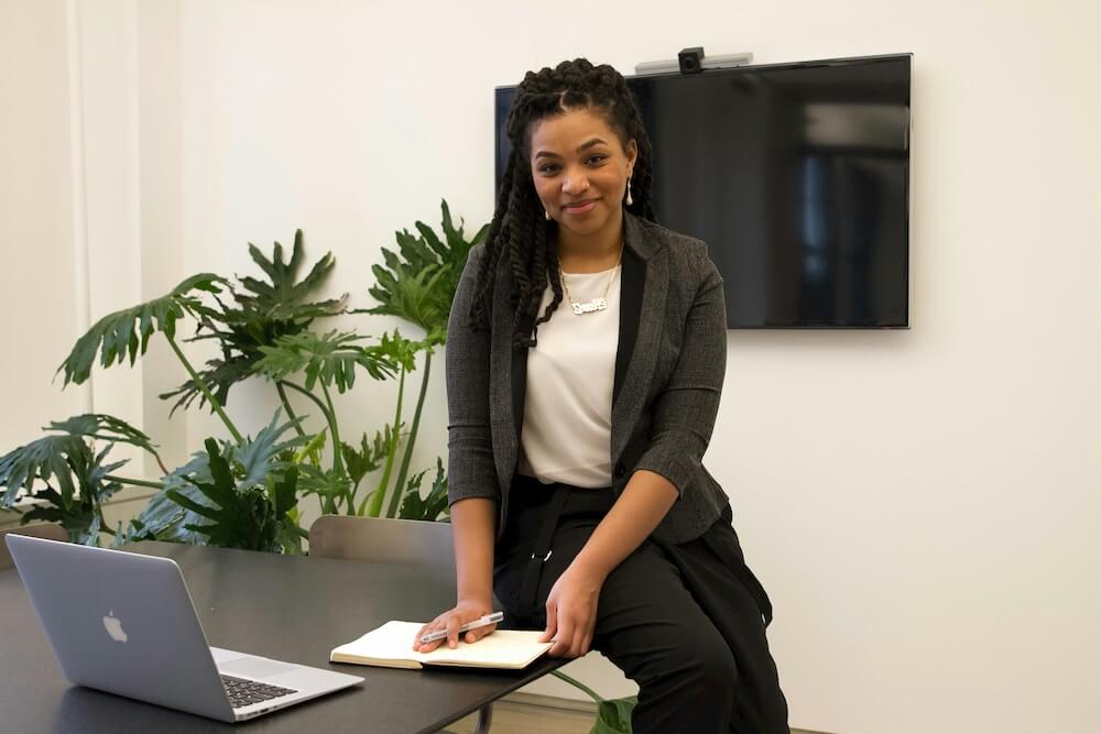 A young Black woman with shoulder-length braids sits casually on the edge of a conference room table, smiling slightly nervously. She wears a modern gray blazer with half-sleeves, a white shirt, and black pants. She holds a pen in one hand and grabs the edge of a notebook with the other. The room is white with a TV and large plant behind her.