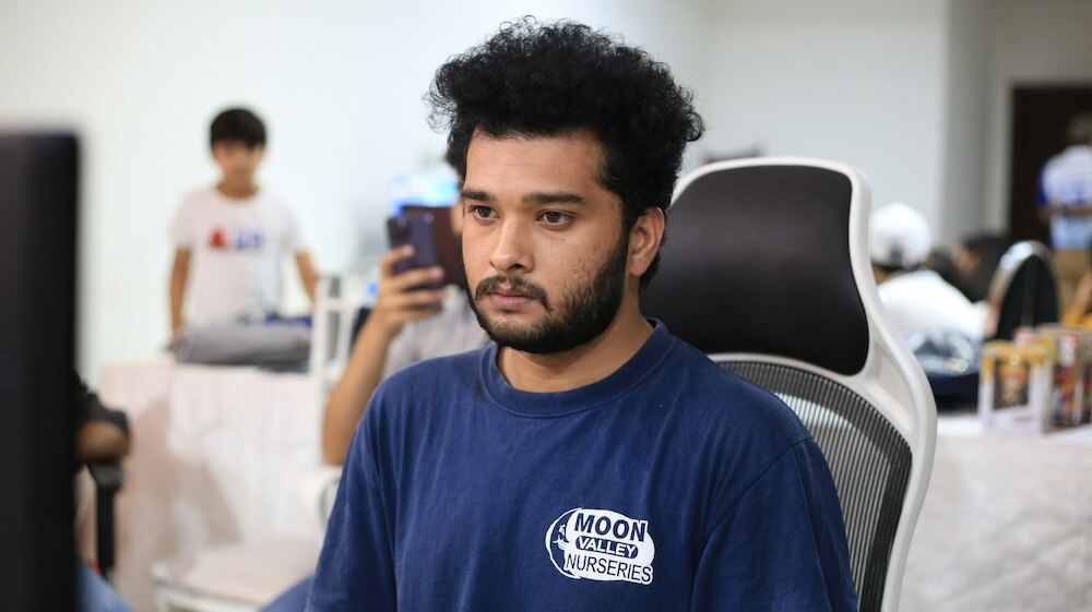 A young man of color wears a blue t-shirt that reads "Moon Valley Nurseries." He sits in a gaming chair, staring blankly at a computer screen in front of him and looking desperately bored