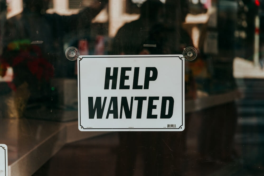 white sign suctioned to a glass window, with black letters in all caps reading "HELP WANTED"