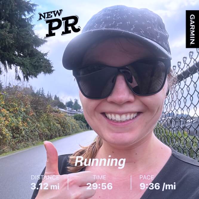 Lucy wears a black baseball hat with silver lightning bolts, black sunglasses, and black longsleeve shirt, pictured from the shoulders up. She smiles widely and gestures with a thumbs-up, the photo adorned with a sticker reading "NEW PR" (personal record) and the word and stats "Running: Distance 3.12 mi Time 29:56 Pace 9:36/mi"