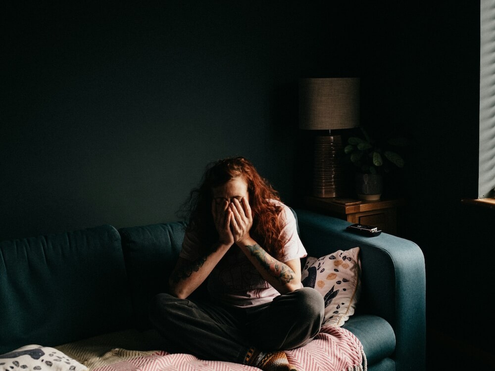 A red haired person with shoulder-length wavy hair and tattoos sits cross-legged on a blue couch in a dark room. Their elbows rest on their knees as they cover their face with their hands, as though upset.