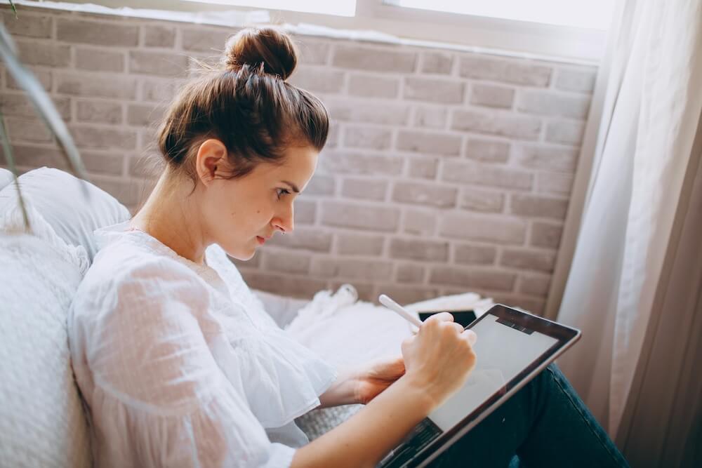A brown-haired white woman with hair in a topknot wears a sheer white blouse and sits on a white couch with legs bent, a tablet resting on her legs. She holds a white stylus in her right hand and is writing or drawing something on the tablet, concentrating.