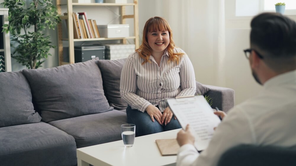 A young white woman with bleached orange wavy shoulder-length hair and straight bangs wears a white and gray vertical striped button-down shirt and jeans. She sits on a gray couch in a casual office setting, hands resting on her legs, as she smiles at a male interviewer across the coffee table from her.