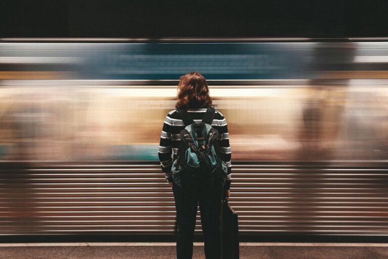 Woman with red hair, black and white striped shirt, black pants, and backpack, seen from the back. She's standing in a subway or train station, with a moving train blurred in front of her