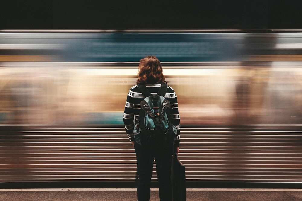 Woman with red hair, black and white striped shirt, black pants, and backpack, seen from the back. She's standing in a subway or train station, with a moving train blurred in front of her