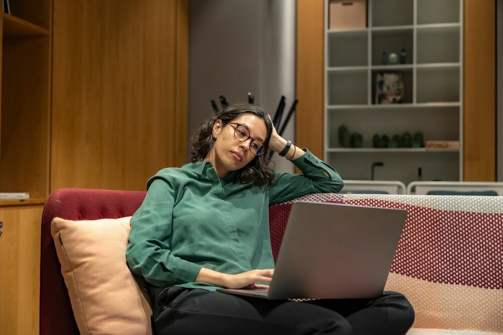A bored feminine-presenting person of color sits casually on a couch, cross-legged with laptop in her lap. She has short black hair and wears a green long-sleeved shirt, black pants, and glasses. She props her head up on one hand, elbow resting on the back of the couch. 