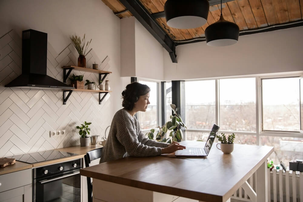 a brunette feminine-presenting person with short hair sits at a large wooden kitchen island, resting arms on the island as she types on her laptop. the room is brightly lit with natural light bouncing off the white walls, and she wears over-the-ear headphones to focus intently on her career change planning.