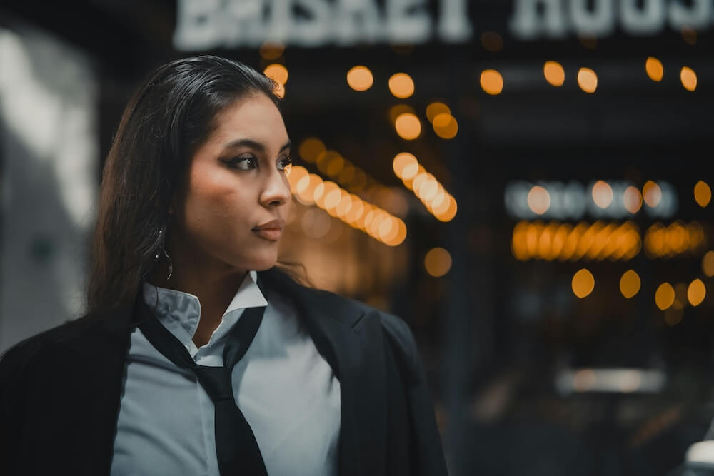 A Latinx feminine-presenting person stands in front of an out-of-focus backdrop of orange lights. They have long black hair and wear a white button-down shirt with collar loosely popped up, a loosened black tie, and a black blazer. They stare off to the side, posed as though lost in thought, but with a confident air.