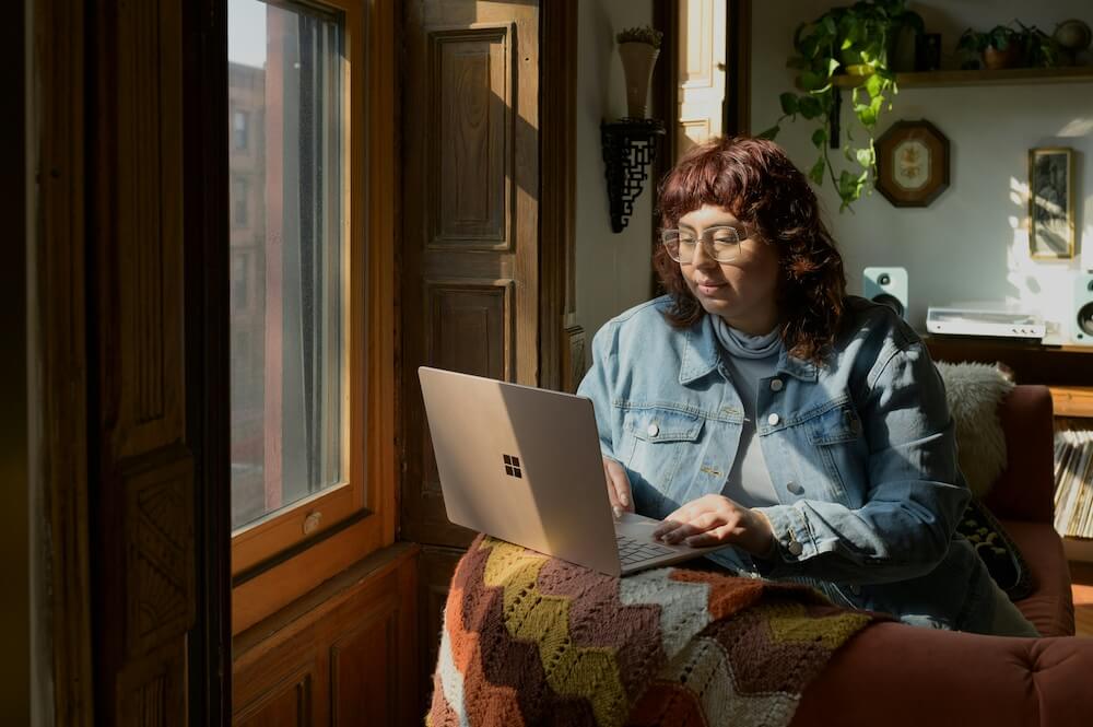 A feminine-presenting person with dyed burgundy hair cut in a bob with microbangs sits on a couch, angled toward a window. She wears a denim jacket and has a laptop rested on the back of the couch, close to the window. She's typing while looking at the screen, and appears to be concentrating but relaxed.