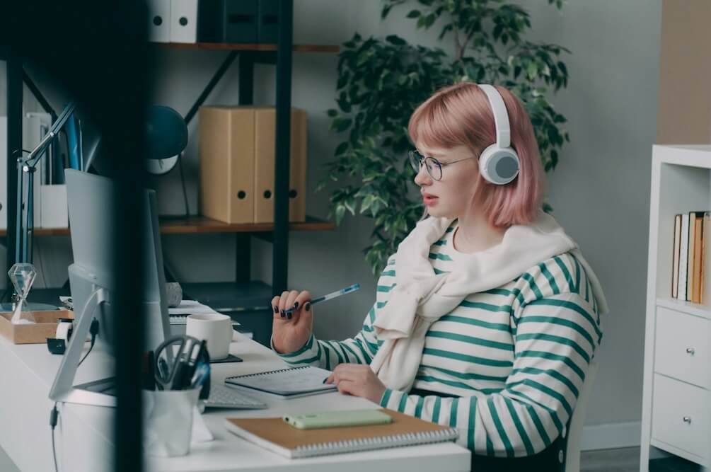 a feminine-presenting person with shoulder-length pink hair wears a green-and-white striped long-sleeved shirt and sits at a white desk in a home office. she might be taking a course about a career she's interested, in a live workshop, or something else, as it looks like she's learning or about to ask a question. her brow is a little furrowed and she raises one hand slightly off the desk, gripping a pen. she may be about to speak, perhaps to ask a question in a workshop or seminar.