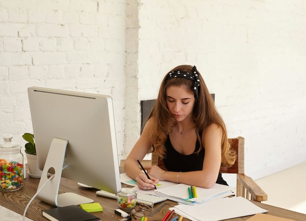 white woman writing in a notebook on a crowded wooden desk in a large office with brick walls painted white. hes desk also has an apple desktop computer, jars of jelly beans, highlighters, and other papers and notebooks.