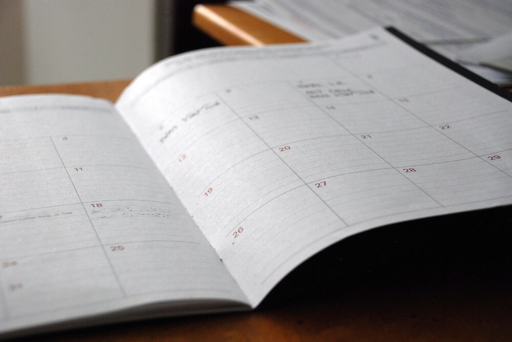 An open paper planner, organized by day of the month, lays on a wood tabletop as though someone is looking at their schedule or about to schedule appointments.